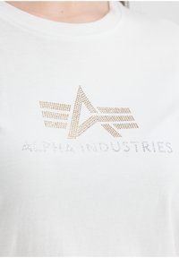 Hvit bomull T-skjorte med et design av gul- og sølvfargede rhinesteiner av Alpha Industries-logoen og et abstrakt stjernegrafikk.