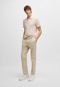 Ανοιχτό ροζ κοντομάνικο πόλο, μπεζ παντελόνι και λευκά αθλητικά παπούτσια. Το πόλο διαθέτει γιακά με δύο κουμπιά και μια διακριτική λεπτομέρεια λογότυπου.