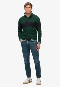 Pullover a righe verde e blu navy con colletto zip, indossato sopra una camicia azzurra chiara, abbinato a jeans blu e sneakers bianche con accenti neri.