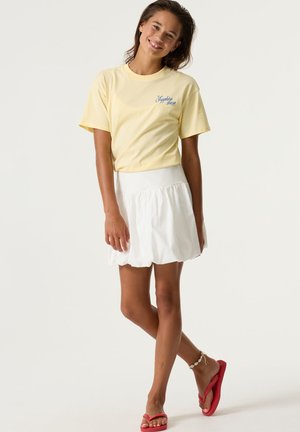 Femme souriante, portant un t-shirt jaune à manches courtes avec le texte « Sapphire shore », une jupe blanche, des tongs rouges et un bracelet de cheville en coquillage, debout jambes croisées.