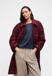 Red and black plaid wool coat with a wide collar, paired with a dark blue top and beige trousers. Features oversized silhouette and pockets.