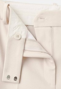 Primo piano della cintura dei pantaloni beige che mostra il bottone, le chiusure a gancio e barra in metallo, la zip e i dettagli del passante per cintura.