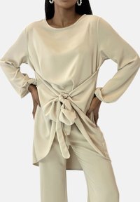 Beige satin-top med rund halsudskæring og lange ærmer. Har en knyttet knudedetalje i taljen og en afslappet pasform. Glat tekstur.