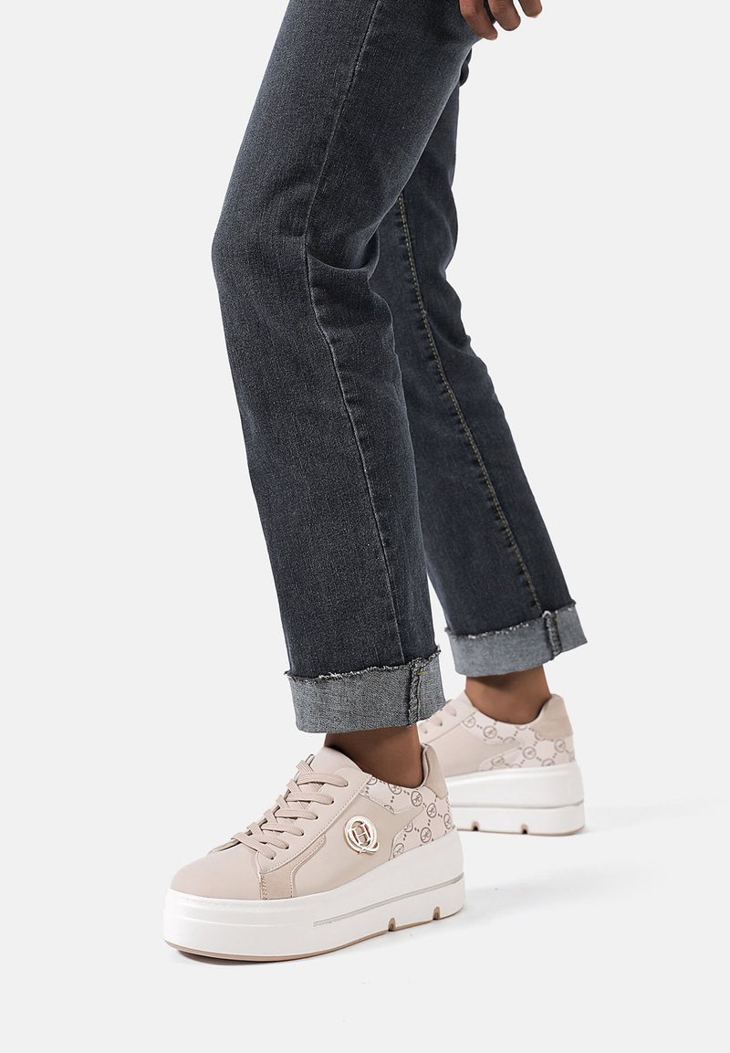 Sneakers platform rosa con dettagli a motivo, dotati di un logo tondo, abbinati a jeans di denim scuri arrotolati. Le sneakers hanno una texture liscia.