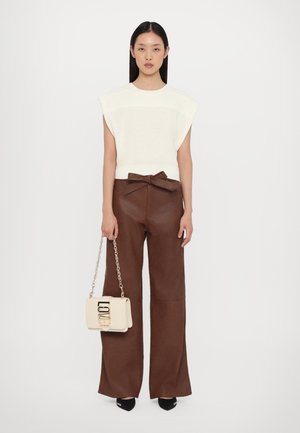 Top a coste color crema con maniche corte, abbinato a pantaloni in pelle marrone a vita alta con fiocco frontale. Borsa leggera beige con tracolla a catena.