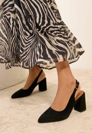 Femeie purtând sandale negre din piele întoarsă cu toc gros și o fustă fluidă cu imprimeu zebră alb-negru, pe un podea cu gresie.