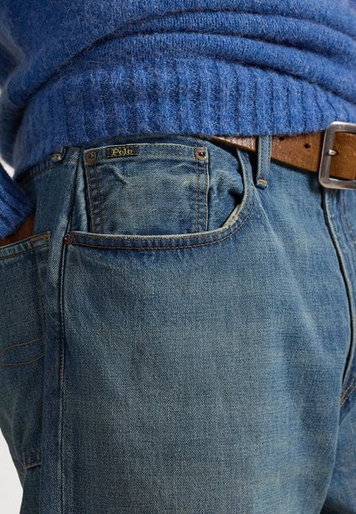 Τζιν παντελόνια σε ανοιχτό γαλάζιο με υφή φινιρίσματος, που διαθέτουν ορατή ετικέτα με λογότυπο και καφέ δερμάτινη ζώνη, αναδεικνύοντας αντίθετα χρώματα και υλικά.