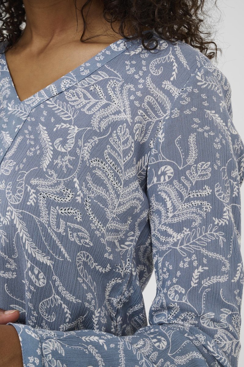 Blaue Bluse mit Muster, die weiße Blumenmotive zeigt, V-Ausschnitt und strukturierter Stoff mit subtilen Rippungen. Lange Ärmel mit Manschetten.