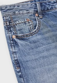 Plavi traper hlače izbliza pokazuju prednji džep, novčanik džepa s metalnim nitnama i vidljive narančaste šavove.