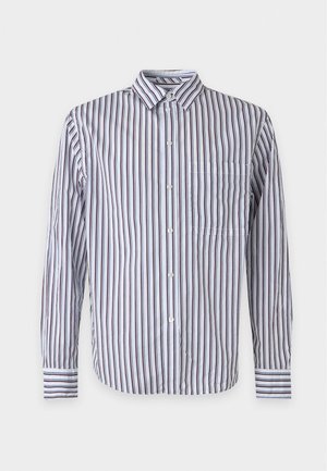 Uomo che indossa una camicia bianca a righe verticali blu navy con bottoni, pantaloni grigio chiaro e scarpe da ginnastica bianche, in piedi su sfondo bianco uniforme.