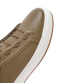 Zapatilla de cuero marrón con una textura suave, que cuenta con cordones en beige, una suela de goma blanca y detalles de costura sutiles.
