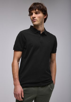 Camisa polo negra con cuello clásico, mangas cortas, tapeta de tres botones y un logo sutil en el pecho. Usada con pantalones verdes oliva.