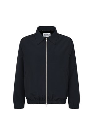 Ung mand iført en sort lynlåstrøje og lyse beige bukser med vidde, står med hænderne i lommerne mod en ensfarvet baggrund.