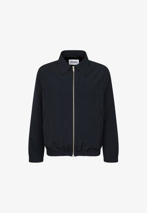 Jonge man met een zwarte jas met rits en lichtbeige wijde broek, staand met handen in de zakken tegen een effen achtergrond.