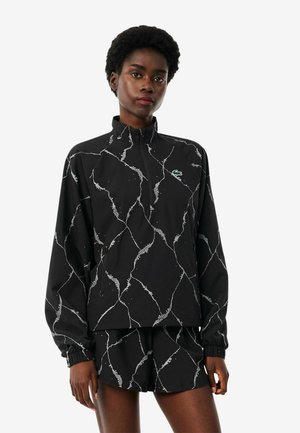 Vrouw draagt een zwart sportjack en korte broek met witte abstracte lijnen en een klein groen logo op de borst van het jack.