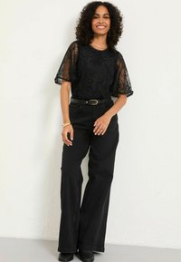 Schwarzes Spitzenoberteil mit durchsichtigen kurzen Ärmeln, kombiniert mit hochtaillierten, schwarzen, weit geschnittenen Hosen und einem schwarzen Gürtel mit goldener Schnalle.