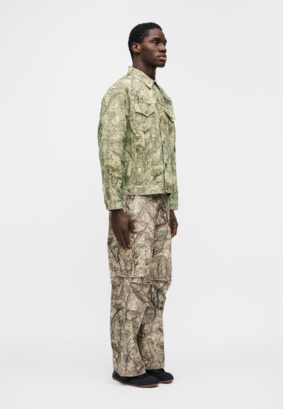 Μπουφάν με σχέδιο καμουφλάζ σε πράσινο χρώμα με τέσσερις τσέπες, συνδυασμένο με παντελόνι καμουφλάζ σε καφέ και πράσινο, φορεμένο με μαύρα παπούτσια.