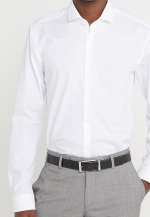 Mężczyzna ubrany w białą, długorękawową koszulę zapinaną na guziki, szare spodnie i czarny pasek z srebrną sprzączką.