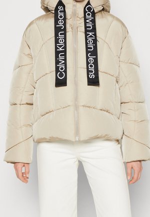 Beżowa pikowana kurtka z kapturem, czarne sznurki z nadrukiem "Calvin Klein Jeans", noszona z białymi spodniami.