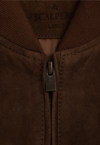 Chaqueta bomber de ante marrón con cuello acanalado, cremallera frontal y detalles cosidos. Etiqueta "Scalpers" visible en el interior.