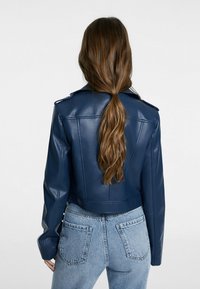 Veste cropped en simili cuir navy avec épaulettes, coupe ajustée et manches longues, assortie à un jean bleu clair. Texture lisse et quincaillerie minimale.