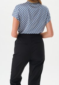 Femme portant un haut à manches courtes à motifs bleu et blanc rentré dans un pantalon noir taille haute, vue de dos.