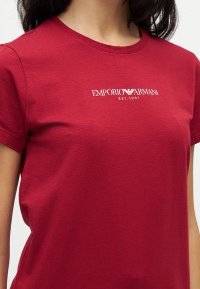 Γυναίκα φοράει κόκκινη μπλούζα Emporio Armani με λευκό λογότυπο και κείμενο "EST. 1981" στο στήθος.