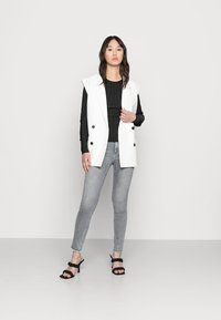 Blazer branco sem mangas com botões pretos, emparelhado com uma blusa preta canelada e jeans cinzentos justos, complementado por sandálias de salto pretas.