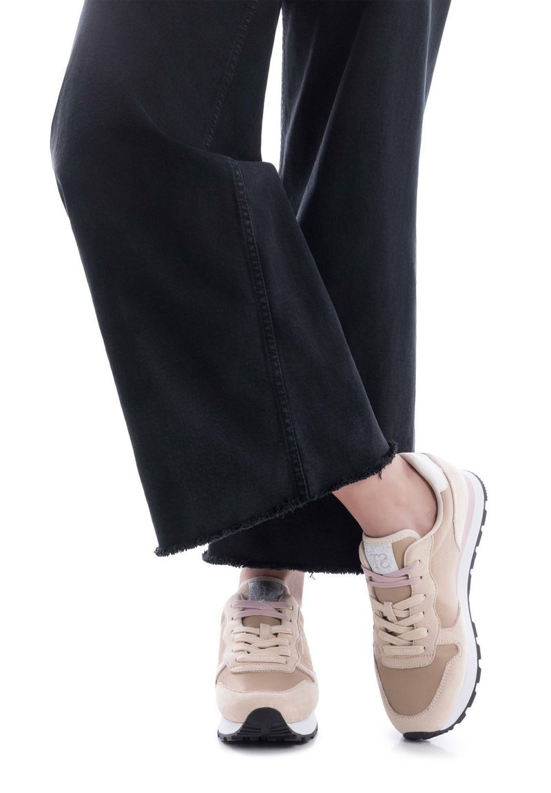 Zapatillas beige de materiales de ante y malla. Cuentan con cierre de cordones, una suela acolchada y un detalle en blanco y rosa en el talón. Se pueden ver jeans negros de pierna ancha.