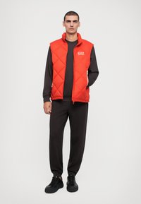 Rød quiltet puffervest med høj krave, lynlåslommer og hvide detaljer, båret over en mørk langærmet skjorte og mørke joggere. Sorte sneakers.
