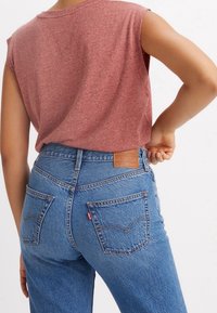 Top sin mangas de brezo rojo combinado con jeans de mezclilla azul de tiro alto que presentan un parche de cuero marrón y costuras clásicas en los bolsillos traseros.