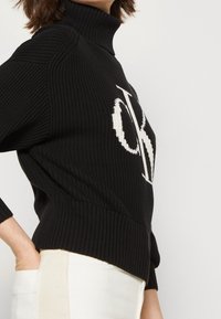 Maglione nero a collo alto lavorato a maglia con un logo bianco sul davanti, texture a coste e una silhouette aderente con maniche a tre quarti.