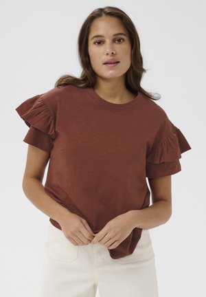 Femme aux longs cheveux bruns portant un haut à manches courtes volantées de couleur rouille et un pantalon blanc taille haute, debout devant un fond uni.