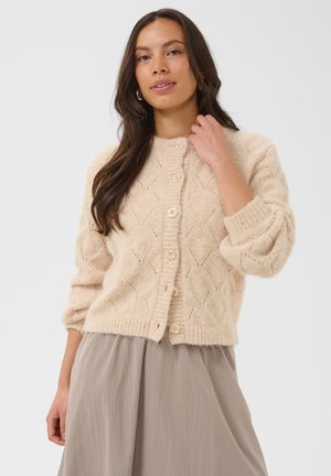 Femme aux longs cheveux bruns portant un cardigan en maille crème à boutons et une jupe taupe, debout devant un fond uni et clair.