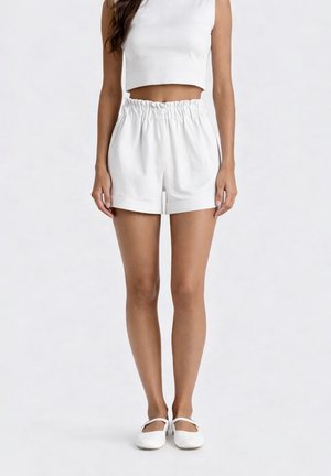 Donna che indossa pantaloncini paperbag bianchi a vita alta, top crop senza maniche bianco e scarpe Mary Jane bianche piatte, in piedi davanti a uno sfondo semplice.