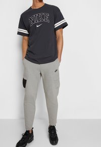 Tmavě šedé tričko Nike s bílými pruhy na krátkých rukávech, spárované se světle šedými tepláky s černou kapsou na cárgo a logem. Černé tenisky.