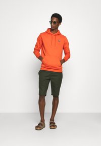 Mężczyzna w pomarańczowej bluzie z kapturem, ciemnozielonych szortach, brązowych sandałach i okularach przeciwsłonecznych stoi z rękami w kieszeni bluzy na białym tle.