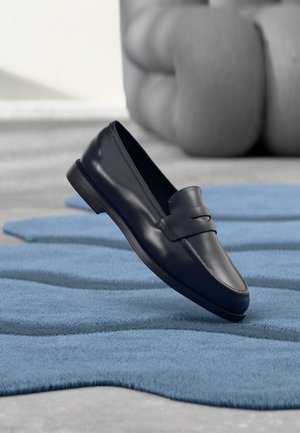 Black leather loafer shoe resting on a textured blue rug with a blurred gray sculpture in the background.