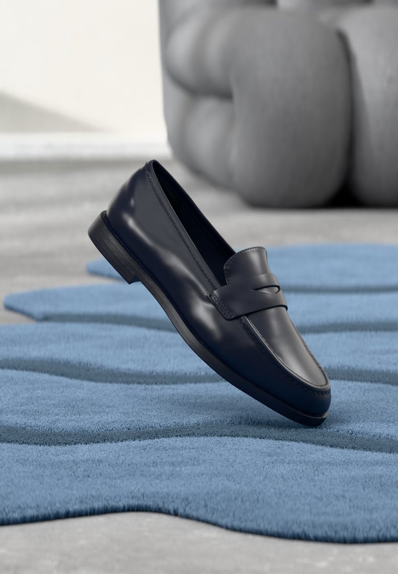 Mocassin en cuir noir posé sur un tapis bleu texturé avec une sculpture grise floue en arrière-plan.