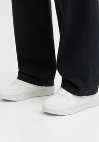 Weiße Ledersneaker mit glatter Textur, runder Spitze und perforierten Details im Obermaterial. Kombiniert mit weit geschnittenen schwarzen Hosen.