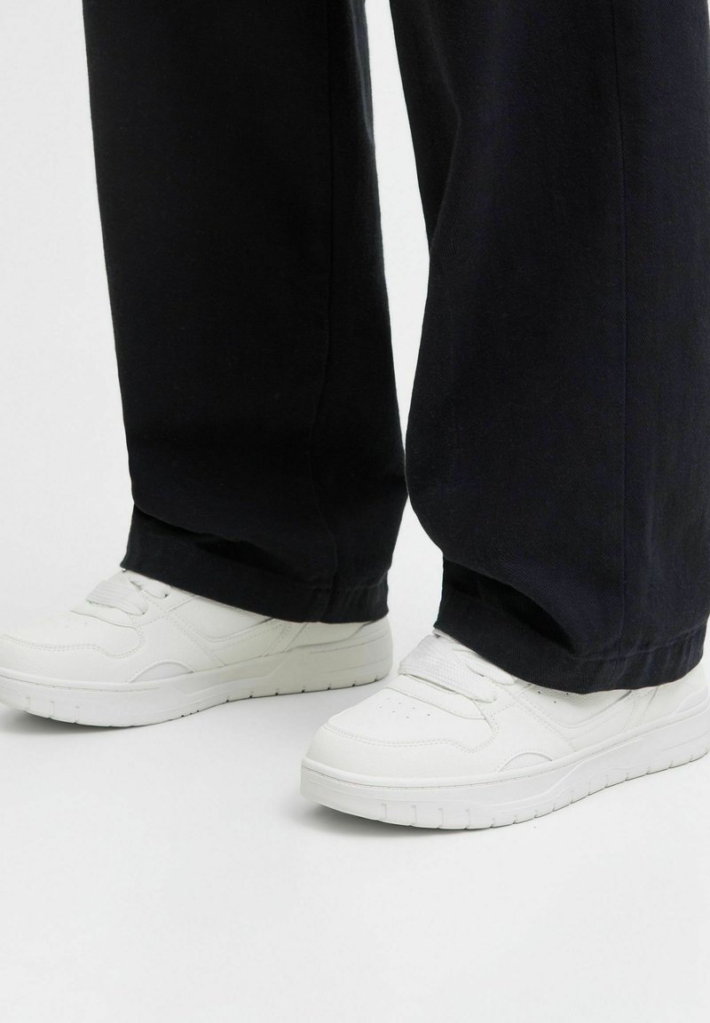 Weiße Ledersneaker mit glatter Textur, runder Spitze und perforierten Details im Obermaterial. Kombiniert mit weit geschnittenen schwarzen Hosen.