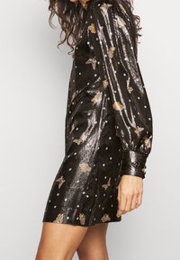 Femme portant une robe noire brillante à manches longues avec des motifs de papillons et des pois blancs, debout devant un fond clair uni.