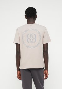 Μπεζ βαμβακερό T-shirt με μεγάλη γκρίζα κυκλική γραφική παράσταση στην πλάτη που αναγράφει "CLOSURE LONDON STUDIOS", κοντά μανίκια και στρογγυλή λαιμόκοψη.