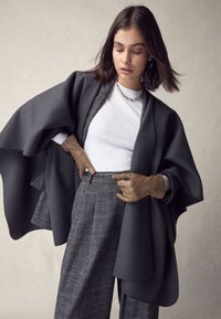 Indumento esterno stratificato grigio scuro con maniche larghe, indossato sopra una maglia bianca a coste, abbinato a pantaloni a quadri strutturati e guanti beige.