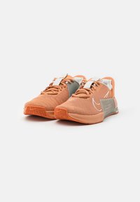 Nike sporta apavi ar sietiņu augšdaļu gaiši oranžā krāsā ar pelēkiem akcentiem, teksturētu gumijas zoli un plakanu šņorējumu drošai piegādei.