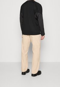 Person wearing black long-sleeve top, beige wide-leg pants with back pocket, and black chunky shoes standing against light gray background.