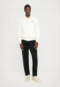 Jeune homme debout, portant un sweat à capuche zippé blanc et un pantalon de jogging noir avec un logo sur la poitrine et la cuisse, baskets blanches, fond neutre.