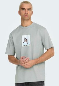 T-shirt di cotone grigio chiaro con una grafica frontale di un canestro da basket e una palla. Maniche corte, scollatura a girocollo e vestibilità rilassata.