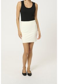 Frau trägt ein schwarzes ärmelloses Top, einen weißen knielangen Rock und schwarze flache Schuhe und steht vor einem einfarbigen Hintergrund.