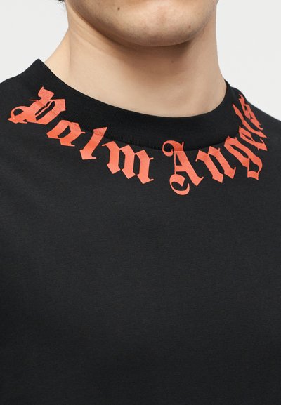 Fekete, kerek nyakú póló, amelynek nyakvonal mentén vörös, gótikus stílusú "Palm Angels" felirat ívelt, egy álltól a felső mellkasig látható személy viseli.